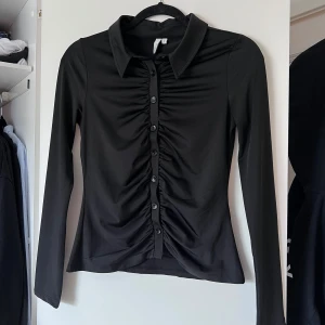 Svart långärmad topp med knappar - Snygg svart långärmad skjorta från Nelly.com med knappar framtill och en stilren krage. Skjortan har en åtsittande passform och är perfekt för en chic look. Tillverkad i ett mjukt och stretchigt material för extra komfort. Använd en handfull gånger 💜 Önskar du veta exakta mått? Tveka inte att höra av dig!