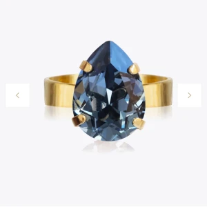 Caroline Svedbom ring - Elegant guldfärgad ring med en stor, droppformad blå sten. Ringen har en justerbar design för att passa olika storlekar. Den är i använt skick men det syns endast på baksidan och är inget man tänker på💕