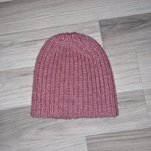 Rosa stickad mössa - Säljer en mysig rosa stickad mössa med ribbad design. Perfekt för kyliga dagar och ger en stilren look. Mössan är mjuk och bekväm att bära. Hemmastickad.