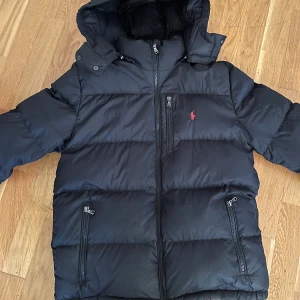 Svart dunjacka från Polo Ralph Lauren - Säljer en svart dunjacka från Polo Ralph Lauren med huva och dragkedja. | Jackan har lite skönhetsfel som har lagats men inget som märks tydligt (se bilder) därav priset, Men annars väldigt bra skick. | Jackan är barnstorlek passar 14 till 16 år. Mellan 160 till 177 cm.