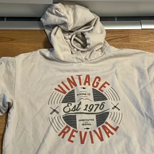 Vit hoodie från ADPT - Säljer en vit hoodie från ADPT med trycket 'Vintage Revival' i rött och svart på framsidan. Hoodien har en klassisk design med dragsko i huvan och är perfekt för en avslappnad stil. Passar bra till jeans eller joggers.