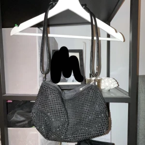 Svart axelväska med stenar  - Snygg svart axelväska täckt med glittrande stenar. Väskan har en dragkedja upptill och en stilren design som passar perfekt för en kväll ute. Den har en lång rem för bekväm bärning över axeln.💞