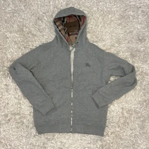 Grå hoodie från Burberry - Säljer en stilren grå hoodie från Burberry med dragkedja och broderad logga på bröstet. Den har en klassisk huva med mönstrat foder och ribbade muddar vid ärmslut och nederkant. Perfekt för en avslappnad look.
