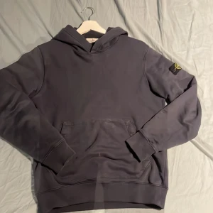 Svart hoodie från Stone Island - Säljer en stilren mörkblå hoodie storlek 14 passar 170 och lite uppåt från Stone Island med deras ikoniska märke på ärmen. Tröjan har en klassisk design med en stor ficka framtill och en bekväm huva. Perfekt för en avslappnad stil. Den har en lite fläk där fram går säkert bort i tvätten. Tvättas innan den fraktats. Obs priset ej hugget i sten.