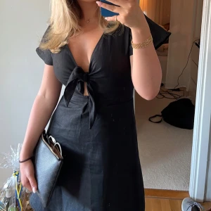 Svart klänning med knyt - Snygg svart klänning med knyt framtill och korta ärmar. Perfekt för en stilren look. Klänningen har en v-ringad design och är gjord i ett lätt material.