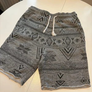 Grå mönstrade shorts från Divided - Snygga grå shorts från Divided med ett coolt geometriskt mönster. De har en bekväm passform med snörning i midjan och är perfekta för en avslappnad stil. Tillverkade i en mjuk bomullsblandning.