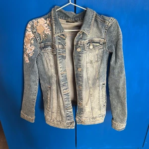 Jeansjacka med blommig brodyr från KappAhl - Snygg jeansjacka från KappAhl i storlek XS. Jackan har en cool blommig brodyr på axeln och klassisk knappstängning framtill.  Jättebra nu till våren. Super snyggt💙🌸