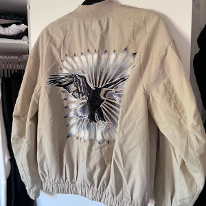 Beige bomberjacka - Säljer en beige bomberjacka från H&M med en cool broderad örn på ryggen. Jackan har dragkedja framtill och två stora fickor. Perfekt för en avslappnad stil. Önskar du veta exakta mått? Tveka inte att höra av dig!