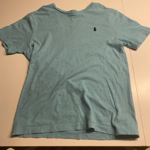 Ljusblå t-shirt från Polo Ralph Lauren - Säljer en ljusblå t-shirt från Polo Ralph Lauren med korta ärmar och en liten broderad logga på bröstet. Perfekt för en avslappnad stil. Tillverkad i mjukt material för extra komfort.