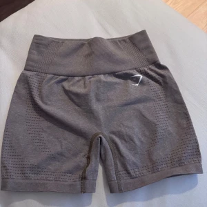 Beige/bruna shorts från Gymshark - Snygga beige shorts från Gymshark med en tight passform. De är tillverkade i ett stretchigt syntetmaterial och har en hög midja för extra komfort. Perfekta för träning och andra aktiviteter. Diskret logga på sidan ger en stilren look. Storlek S.