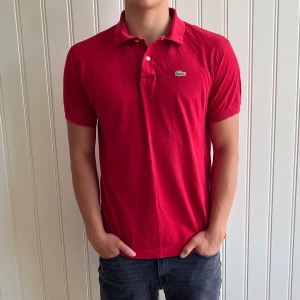 Lacoste Piké - Säljer en snygg röd pikétröja från Lacoste. Den har en klassisk krage med två knappar och det ikoniska krokodilmärket på bröstet. Passar storlek M. 