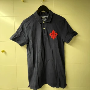 Mörkblå pikétröja från Morris - Snygg mörkblå pikétröja från Morris med röd dekorativ detalj på bröstet och text på ryggen. Tröjan har korta ärmar och klassisk krage med knappar. Perfekt för en stilren look.