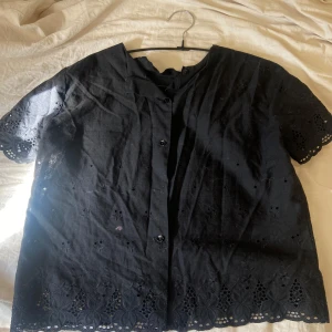 Svart kortärmad blus med broderi - Bra skick förutom att den saknar en knapp. Svart skjorta i bomull med korta ärmar och fina broderade detaljer längs ärmslut och nederkant. Skjortan har knappar framtill och ett luftigt, genombrutet mönster som ger en cool och somrig känsla. Perfekt för dig som gillar unika detaljer och stilren look.
