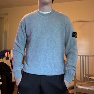 Grå Stone Island sweatshirt i bomull - Grå sweatshirt från Stone Island med klassisk rund hals och ikonisk patch på vänster ärm. Tröjan är i mjuk bomull och har en relaxed passform med långa ärmar och ribbade muddar. Perfekt för dig som gillar stilren streetwear. Ett litet hål som man ser på sista bilden 