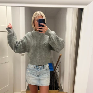 Grå stickad croppad tröja - Supermysig grå/ grön stickad tröja med croppad passform och breda, långa ärmar. Tröjan har ribbad struktur och en högre rund halsringning. Perfekt att styla med jeans eller kjol för en chill look.