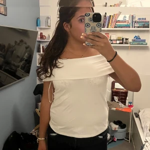 Vit offshoulder topp i satin - Snygg vit offshoulder topp i satin med bred krage som vilar över axlarna. Toppens släta material ger en lyxig känsla och den är perfekt för dig som vill ha en stilren och trendig look. Passar till både jeans och kjol för en cool vibe.