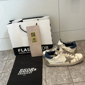 Golden Goose vita sneakers med blå detalj - Golden Goose sneakers i vitt skinn med grå snörning, grå stjärndetalj på sidan och blå hälkappa. Klassisk låg modell med rund tå och platt sula. Snyggt slitna för en cool, avslappnad vibe. Kommer med originalpåse och förpackning.