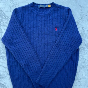 Blå stickad tröja från Polo Ralph Lauren - Säljer en mysig blå stickad tröja från Polo Ralph Lauren med klassiskt kabelstickat mönster och röd broderad logga på bröstet i storlek M. Jag köpte den nyskriven av en annan person men säljer den vidare då den tyvärr inte passade mig. Tröjan är i nyskick. 