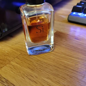 Giorgio Armani Sì parfym - Elegant parfymflaska från Giorgio Armani Sì med stilren, genomskinlig glasdesign och svart kork. Innehållet är bärnstensfärgat och flaskan har märkets logga tryckt framtill. Perfekt för dig som gillar lyxiga dofter.