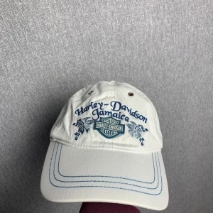 Vit Harley-Davidson keps Jamaica - Vit keps från Harley-Davidson med Jamaica-brodyr i blått framtill och klassisk logga. Justerbart spänne bak i metall och blå dekorativa sömmar på skärmen. Tillverkad i bomull. Liten fläck går att fixa