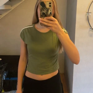 Grön croppad topp  - Säljer en grön croppad topp från FB Sister med vita ränder på axlarna. T-shirten har rund halsringning, korta ärmar och är i mjuk bomull. Perfekt för dig som gillar sportig och avslappnad stil.💚