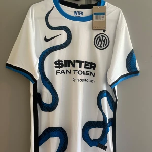 Inter Milan vit bortatröja Nike - Inter Milans officiella bortatröja från Nike i vitt med svart och blått ormmönster över framsidan. Tröjan har klubbmärke, Nike-logga och sponsortryck. Rund hals med blå kant och svarta detaljer på ärmarna. Tillverkad i lätt och ventilerande material.