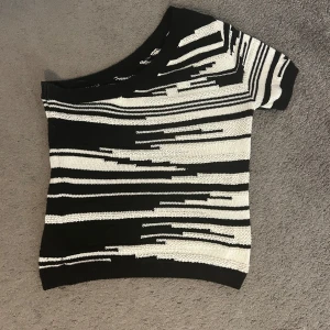 Svartvit offshoulder stickad topp - Säljer en unik stickad topp i svart och vitt med coolt randigt mönster. Toppen har en offshoulder-design och kort ärm, vilket ger en trendig och avslappnad vibe. Perfekt för dig som gillar att sticka ut med grafiska detaljer och sköna material.