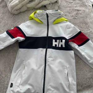 Helly Hansen vit vindjacka med huva - Snygg vit vindjacka från Helly Hansen med svart och röd detalj på ärmarna samt en gul huva. Jackan har dragkedja framtill, två fickor med dragkedja och HH-logga på bröstet. Perfekt för blåsiga dagar och streetstyle.
