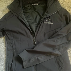 Arcteryx windbreaker - Säljer en svart vindjacka från Arc'teryx i storlek XS. Jackan har dragkedja framtill, hög krage och diskret logga på bröstet. Perfekt för blåsiga dagar och outdoor-aktiviteter. Snygg och stilren design som funkar till det mesta.