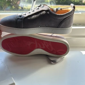 Grå mocka sneakers från Louboutin - Snygga grå sneakers i mocka från Christian Louboutin med klassisk röd sula och vita kanter. Använda ett par gånger men fortfarande i bra skick går att göra de snyggare med lite sko tvätt kit så blir skorna mycket snyggare. Skorna har rund tå, snörning och diskreta metalldetaljer vid sidan. Insidan är fodrad i ljust skinn för extra komfort. Perfekt för dig som vill sticka ut med exklusiva detaljer.