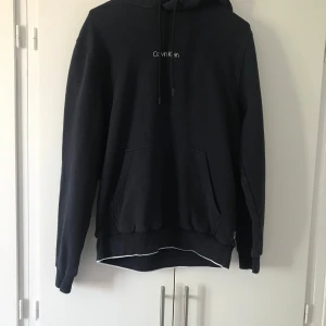 Calvin klein Hoodie  - Mörkblå hoodie från Calvin Klein med vit logga på bröstet. Klassisk känguruficka framtill, huva med snörning och ribbade muddar. Perfekt för en chill och stilren look. Mjuk och skön bomullsblandning, passar dig som gillar enkel och clean streetstyle.
