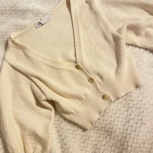 Beige stickad kofta från Zara - Superfin beige stickad kofta från Zara med v-ringning och tre knappar framtill. Modellen är croppad med bred ribbad mudd i midjan och vid ärmslut. Perfekt att slänga över en topp för en chill och trendig look.