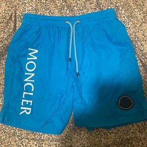 Blå Moncl3r badshorts med snörning - Snygga blå badshorts från Moncl3r med vit logga längs benet och klassisk Moncler-patch. Shortsen har elastisk midja med snörning och innerbyxa i mesh. Perfekta för stranden eller poolen, riktigt fräsch färg och exklusiv känsla. Använda 1 gång som nya 