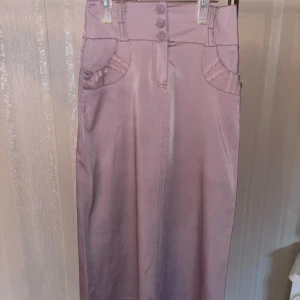 Ljuslila långkjol med dekorativa knappar och brodering  - Långkjol i ljuslila med snygga dekorativa knappar framtill och coola fickdetaljer samt brodering. Kjolen har hög midja, bälteshällor och raka linjer. Materialet är slätt och känns som bomull eller polyester. Perfekt för dig som vill sticka ut med en unik och trendig look.(Står 38 men den är absolut inte det, den är 36/34)