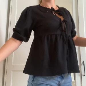Svart blus med knyt och puffärm - Supersöt svart blus med puffiga halvlånga ärmar och knytning fram i halse. Perfekt för dig som har en typ av Stockholmsstil. Blusen har en lätt rynkad midja som ger en flowy och avslappnad siluett. Perfekt till jeans eller kjol.  Aldrig använd!