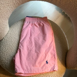 Rosa badshorts från Polo Ralph Lauren - Snygga rosa badshorts från Polo Ralph Lauren med elastisk midja och klassisk blå logga broderad framtill. Shortsen har bakficka med lock och innerfoder i mesh. Perfekta för sommardagar vid stranden eller poolen. Har ett par andra inne för samma pris. 2 För 1000/ 1 för 599 