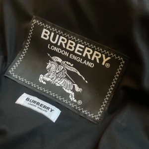 Svart jacka från Burberry - Snygg svart T-Shirt från Burberry London England med klassisk logga och detaljerad brodyr på insidan. Jackan har en stilren look och känns lyxig, perfekt för dig som gillar exklusiva märken och tidlös design.