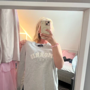 Grå stickad tröja från Morris Lady - Superfin ljusgrå stickad tröja från Morris Lady med broderad logga framtill. Klassisk rund halsringning och långa ärmar. Perfekt till jeans eller kjol för en clean och stilren look.