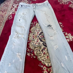 Dolce & Gabbana låga bootcut jeans - Säljer ett par ljusblå lågmidjade jeans från Dolce & Gabbana med slitningar och hål på benen. Bakfickan har D&G-logga i svarta nitar. Jeansen har klassisk femficksmodell och raka ben. Perfekta för dig som gillar en edgy och trendig look. Står ingen storlek, skulle tippa på xs,s. (Skriv för mått) 