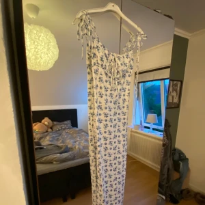 Vit långklänning med blå blommor - Supersöt långklänning i vitt med blått blommönster. Klänningen har smala axelband med knyt och en söt snörning framtill vid bysten. Perfekt för sommarens alla tillfällen och ger en romantisk vibe.