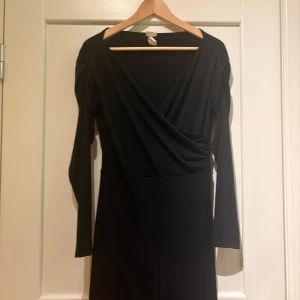 Svart jumpsuit med långa ärmar - Stilren svart jumpsuit med omlott-skärning och långa ärmar. Axelpartiet har lätt rynkade detaljer som ger en snygg touch. Jumpsuiten har en djup v-ringning och är figurnära upptill. Perfekt för dig som gillar enkel men elegant stil.
