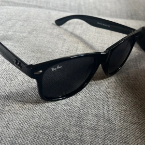 Svarta Ray-Ban Wayfarer solglasögon - Klassiska svarta Ray-Ban Wayfarer solglasögon med glansig finish och ikonisk fyrkantig form. Snygg logga på bågarna och mörka linser. Tillverkade i Italien och passar perfekt för en stilren look. Kan gå ner i pris vid snabb affär