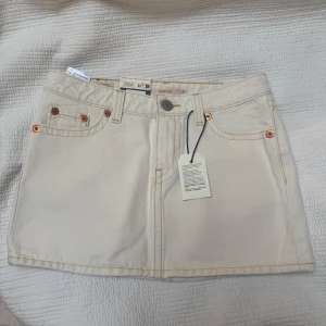 Vit jeanskjol från Levi's - Snygg vit jeanskjol från Levi's med klassiska fem fickor, synliga sömmar och detaljer i kopparfärg. Storlek 25, aldrig använd med tagg på.