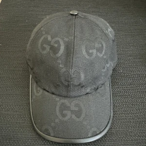 Svart keps från Gucci med GG-mönster - Snygg svart keps från Gucci med ikoniskt GG-mönster över hela kepsen. Kepsen har böjd skärm med svart kant i skinn och justerbart band bak. Tillverkad i bomullsmix med exklusiv känsla och diskret logotyp på insidan.