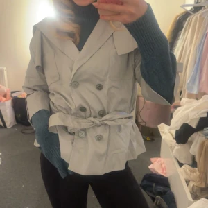Ljusgrå trenchcoat med bälte - Snygg ljusgrå trenchcoat med dubbelknäppning och knytskärp i midjan. Jackan har bred krage och trekvartsärmar, vilket ger en cool och modern vibe. Perfekt för dig som vill ha en stilren och trendig jacka till vår eller höst.
