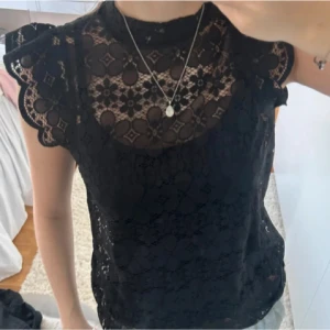 Svart spetsblus med volangärmar - Supersnygg svart blus i spets med blommigt mönster och volangdetaljer vid axlarna. Hög hals och ärmlös design ger en chic vibe. Perfekt att styla med jeans eller kjol för en trendig look.