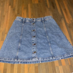 Blå jeanskjol från H&M Divided - Snygg blå jeanskjol från H&M Divided med knappar hela vägen framtill och bälteshällor i midjan. Kjolen har en klassisk A-linjeform och är i kort modell, perfekt till sneakers eller boots.