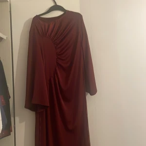 Vinröd långklänning med drapering - Säljer en vinröd långklänning med snygg drapering framtill och vida ärmar. Klänningen har en elegant, flowy passform och är tillverkad i ett glansigt material som ger en lyxig känsla. Perfekt för dig som vill sticka ut med en unik och stilren look. Passar bra för S/M. Använt en gång i bröllop. 