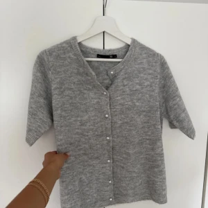 Grå kofta med pärlknappar från 157 - Supermjuk grå kofta från 157 med kort ärm och snygga pärlknappar framtill. Koftan är stickad och har en klassisk rund halsringning. Perfekt att slänga över en topp för en chill look. Aldrig använd