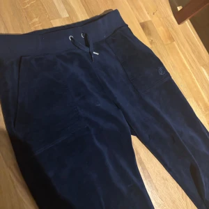 Mörkblå velourbyxor Juicy Couture XS - Mörkblå mjukisbyxor från Juicy Couture i lyxig velour. Byxorna har resårmidja med snörning och sidofickor, samt en diskret logga broderad på ena benet. Perfekta för en chill och trendig look.